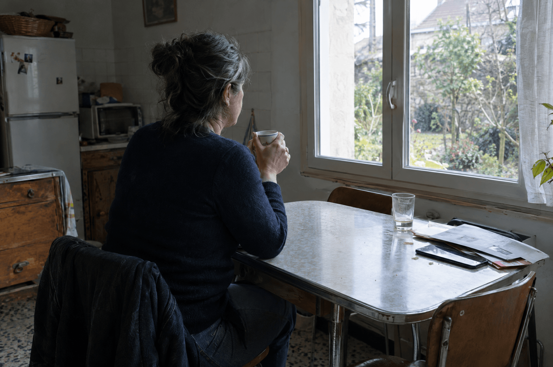 Une femme de dos à une table de cuisine, une tasse dans les mains.
