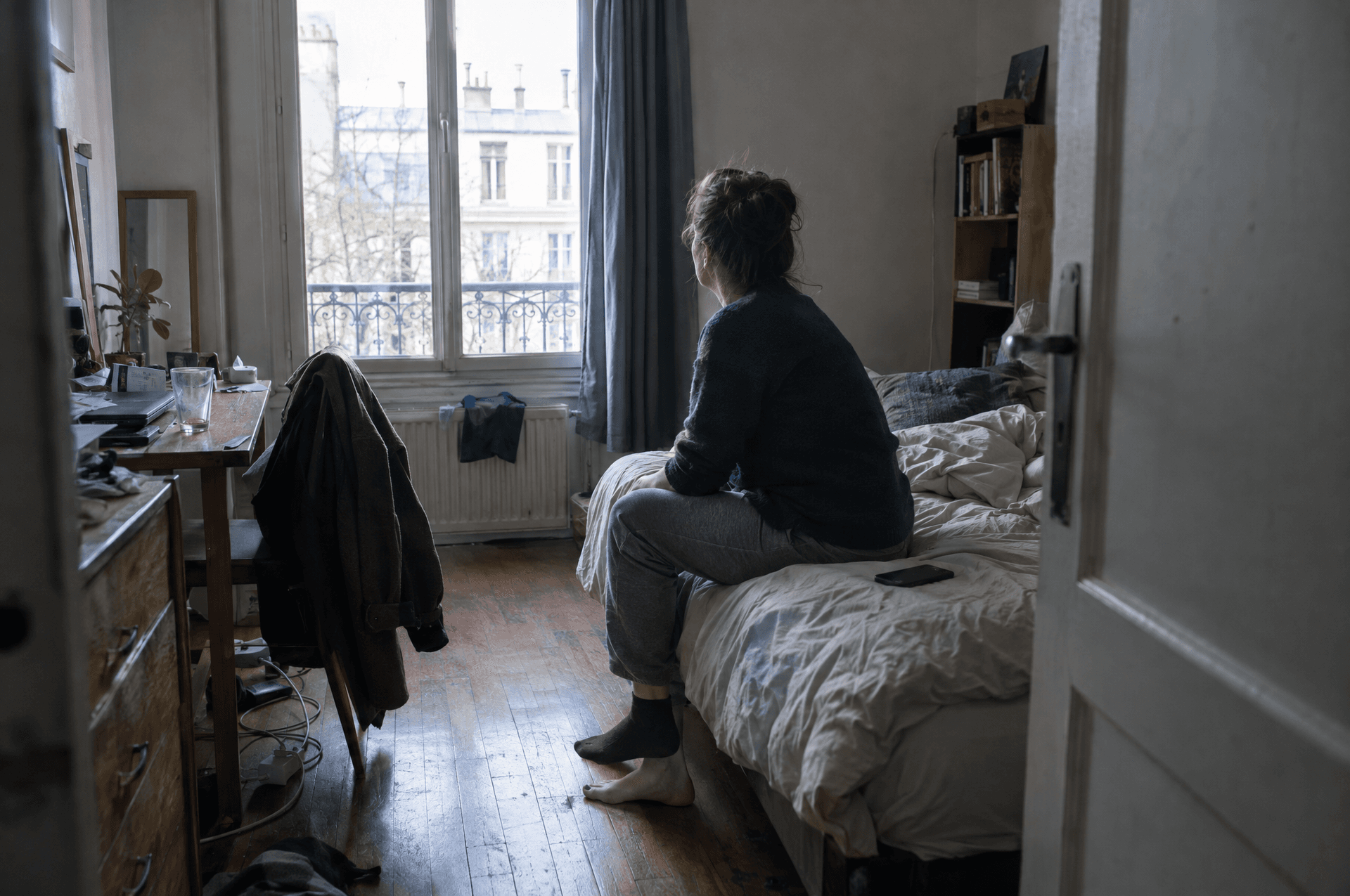 Une femme assise au bord de son lit le matin, le regard tourné vers la fenêtre.