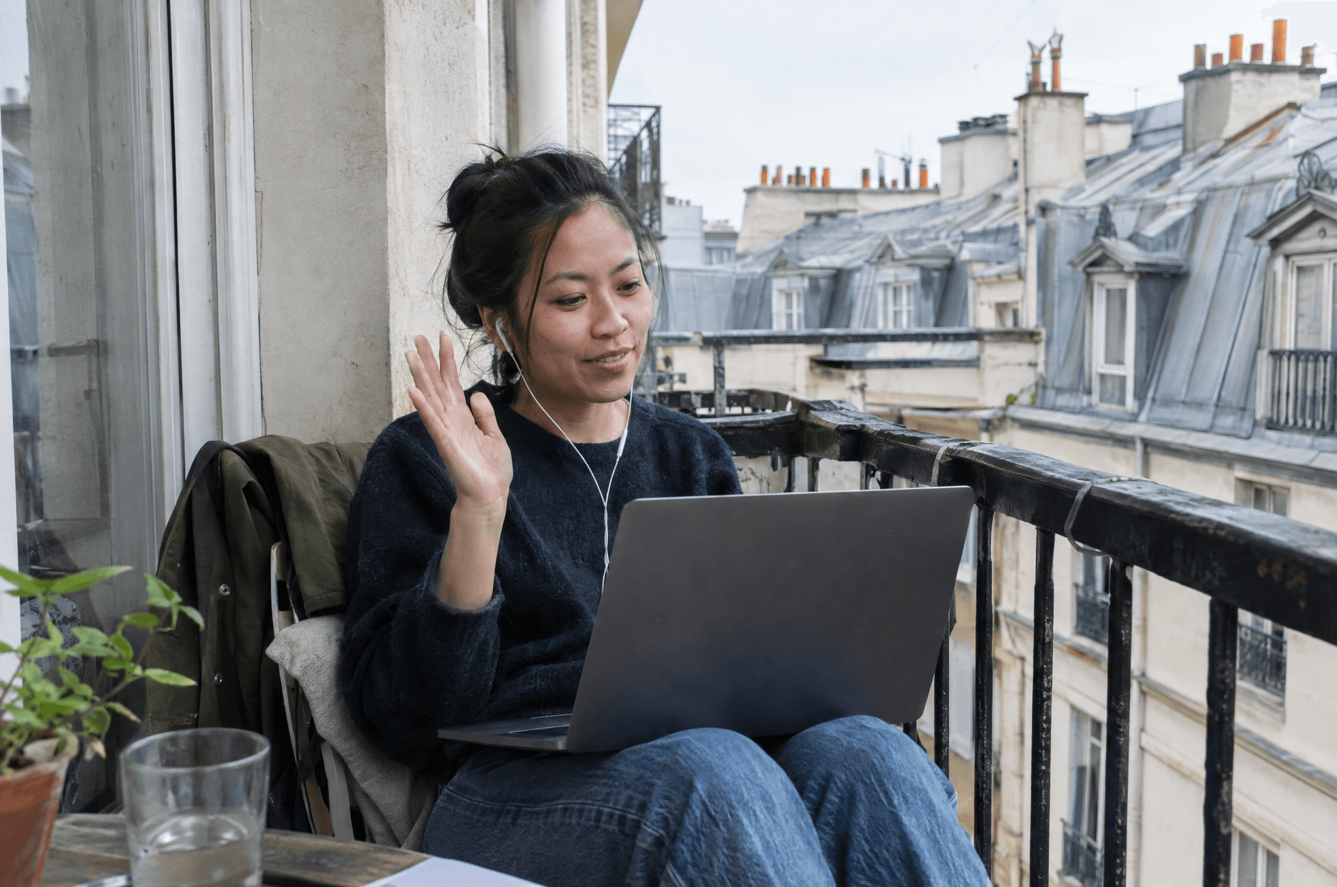 Une femme sur un balcon, un ordinateur sur les genoux, qui fait un petit signe de la main.